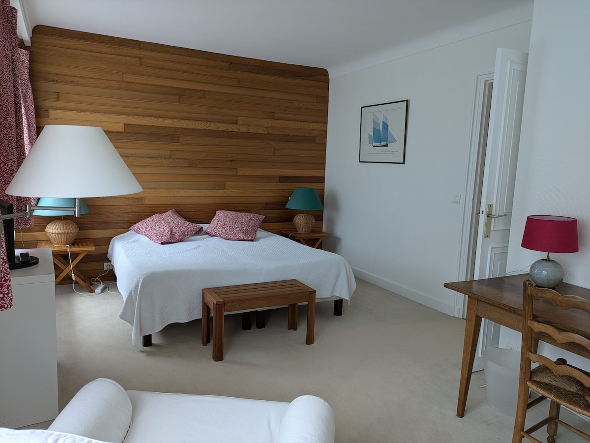 A spacious room is presented with a large bed featuring two decorative pillows. Natural wood paneling adorns one wall, while a simple wooden bench and a chair complete the furnishings. Soft lighting is provided by a floor lamp and bedside lamps.