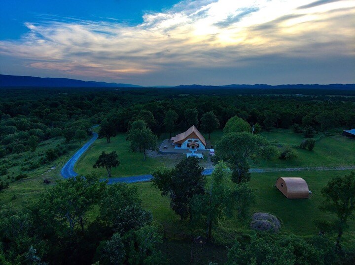 An aerial view of the two-level cabin surrounded by lush green trees and expansive natural landscape. A winding dirt road leads to the cabin, set against a backdrop of rolling hills and a colorful sky at dusk.