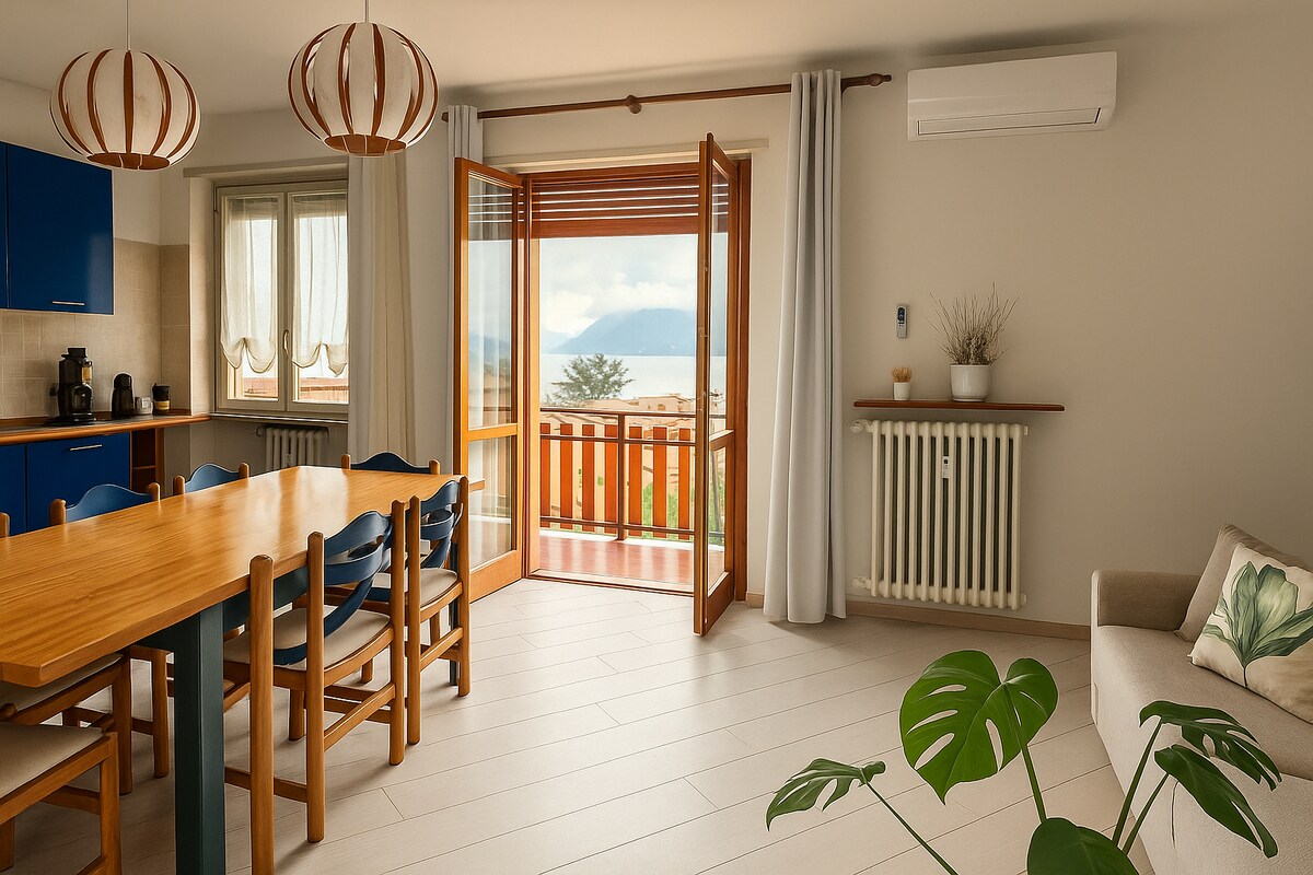 The living area features a wooden dining table surrounded by blue chairs. Large windows and double doors open to a view of the lake, allowing natural light to fill the space. A cozy sofa sits against the wall, complemented by decorative plants.