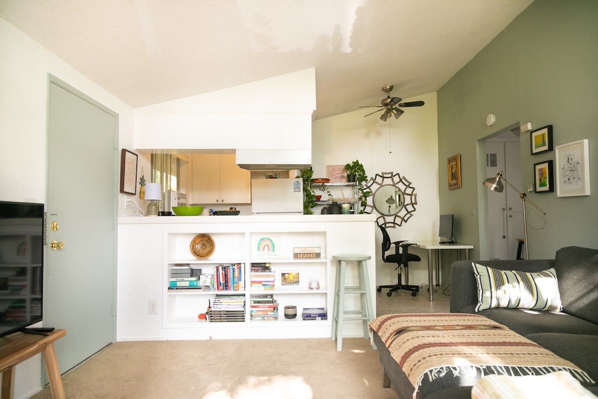 An open living area is presented with a cozy sofa beside a light-colored coffee table. A kitchen bar with white cabinetry and a plant is visible in the background. A designated workspace with a desk and chair is positioned for convenience. Bookshelves display various items, adding character.