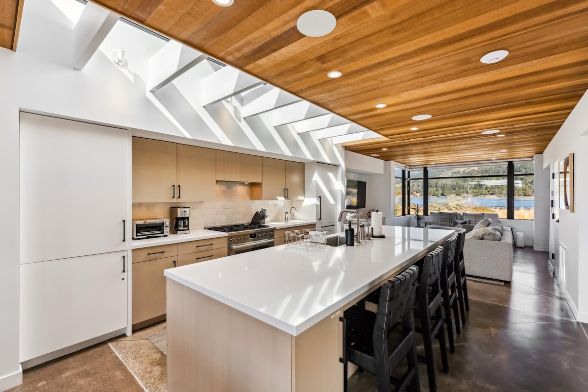 A modern kitchen design features sleek cabinetry and an expansive island with seating for multiple guests. Large windows allow natural light to fill the space, complemented by a wood-paneled ceiling that adds warmth to the contemporary aesthetic.