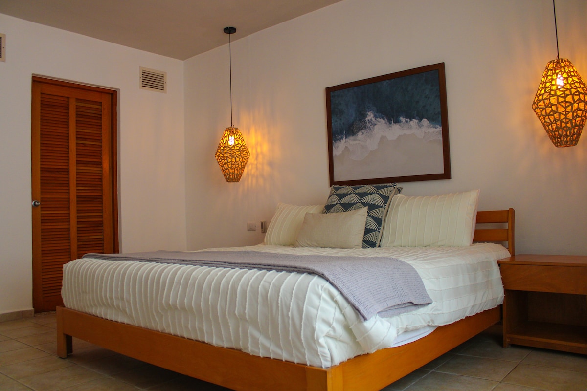 A spacious bedroom features a large bed with soft linens and decorative pillows. Soft lighting is provided by woven pendant lamps, casting a warm glow. A piece of art depicting ocean waves hangs on the wall, and a wooden nightstand is positioned beside the bed.
