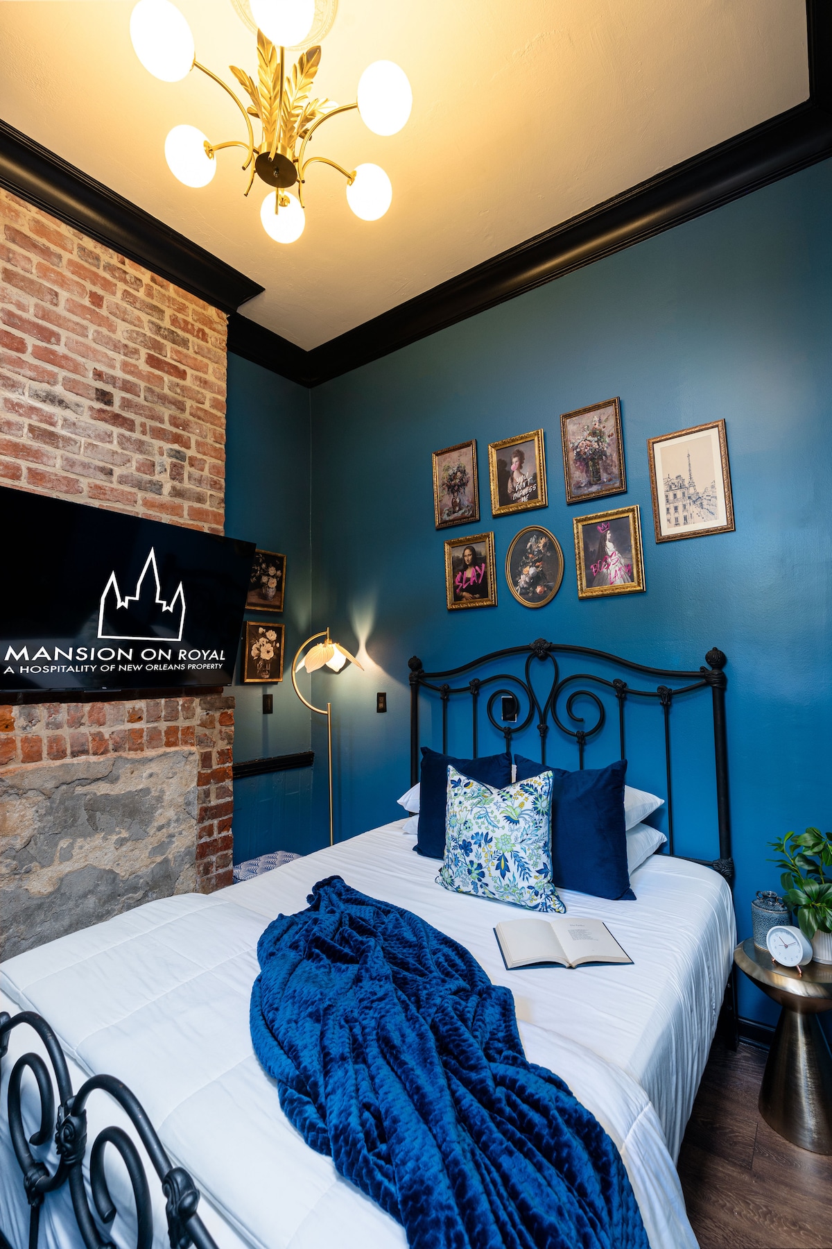 The bedroom features a wrought-iron bed adorned with plush pillows and a deep blue throw blanket. A textured brick wall lends character, while framed artwork decorates the blue walls. A flat-screen TV is mounted nearby, and an open book rests on the bed.