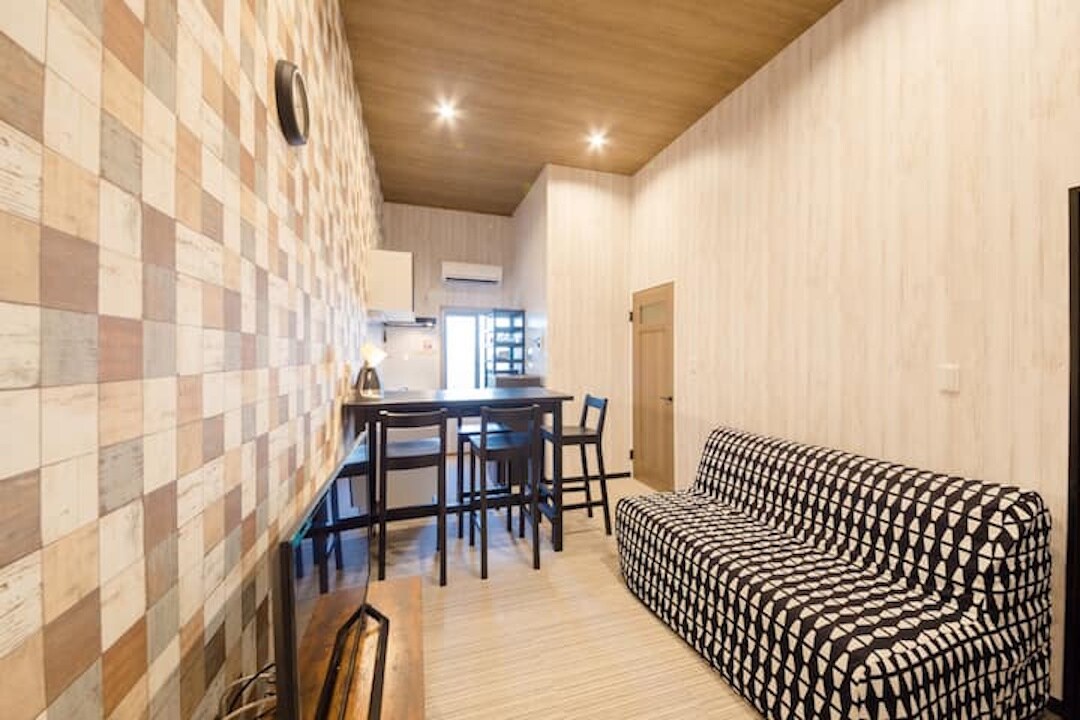 A stylish living area features patterned seating along one wall, complemented by a contrasting wooden ceiling. A dining area with dark high chairs is positioned against the adjacent wall, with the kitchen area visible in the background.