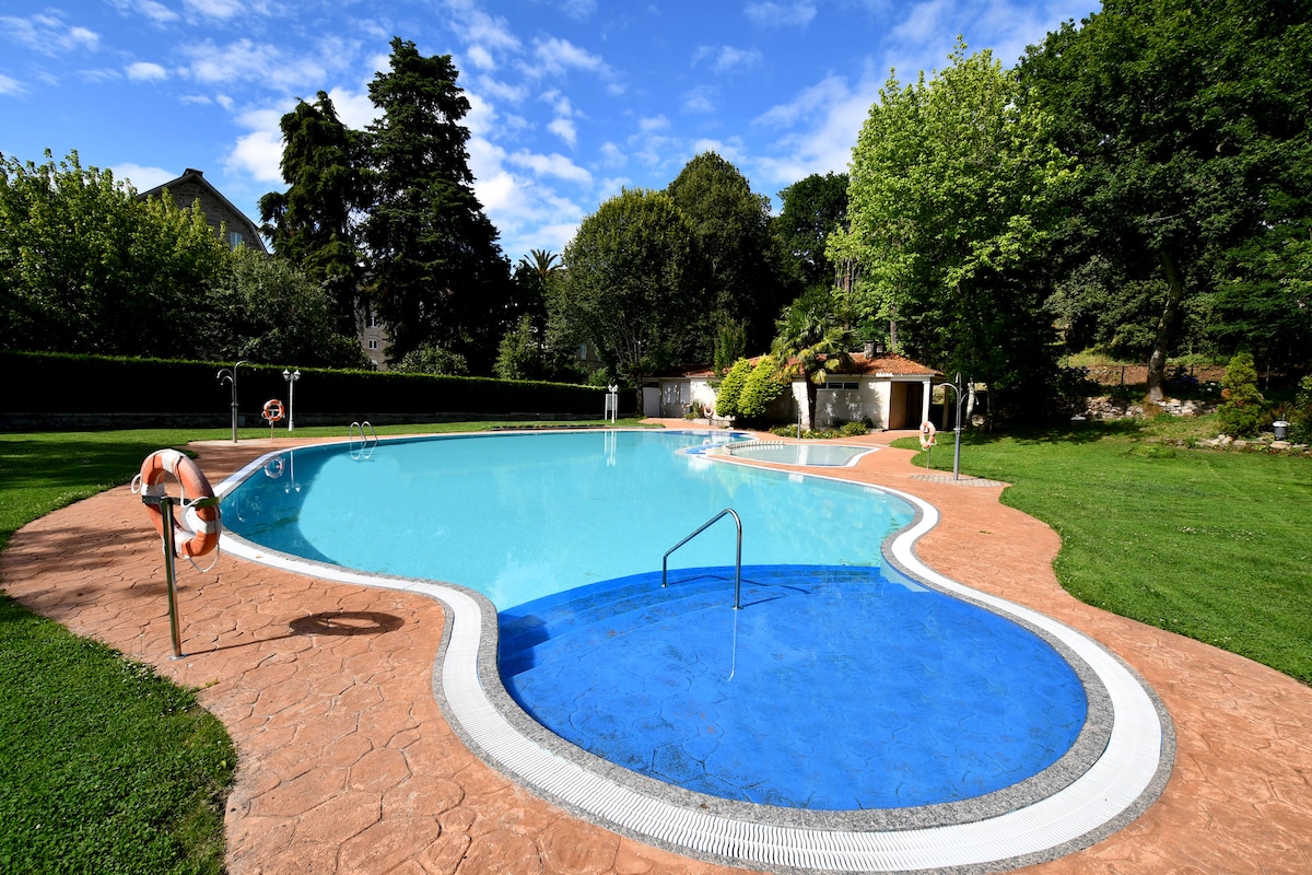 The image showcases a spacious swimming pool surrounded by lush greenery and trees. The pool's unique shape includes shallow and deep areas, with steps leading into the water. A grassy area borders the pool, accompanied by a relaxing ambiance created by the natural surroundings.