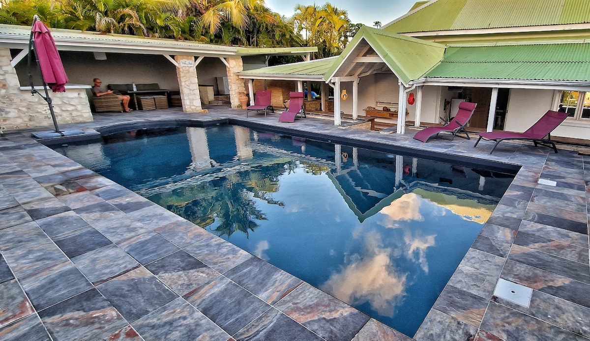 A saltwater pool is centered within a patterned stone deck, reflecting the sky and surrounding palm trees. Nearby shaded seating areas are visible, along with lounge chairs positioned for relaxation. The pool area is complemented by charming architectural elements of the villa.
