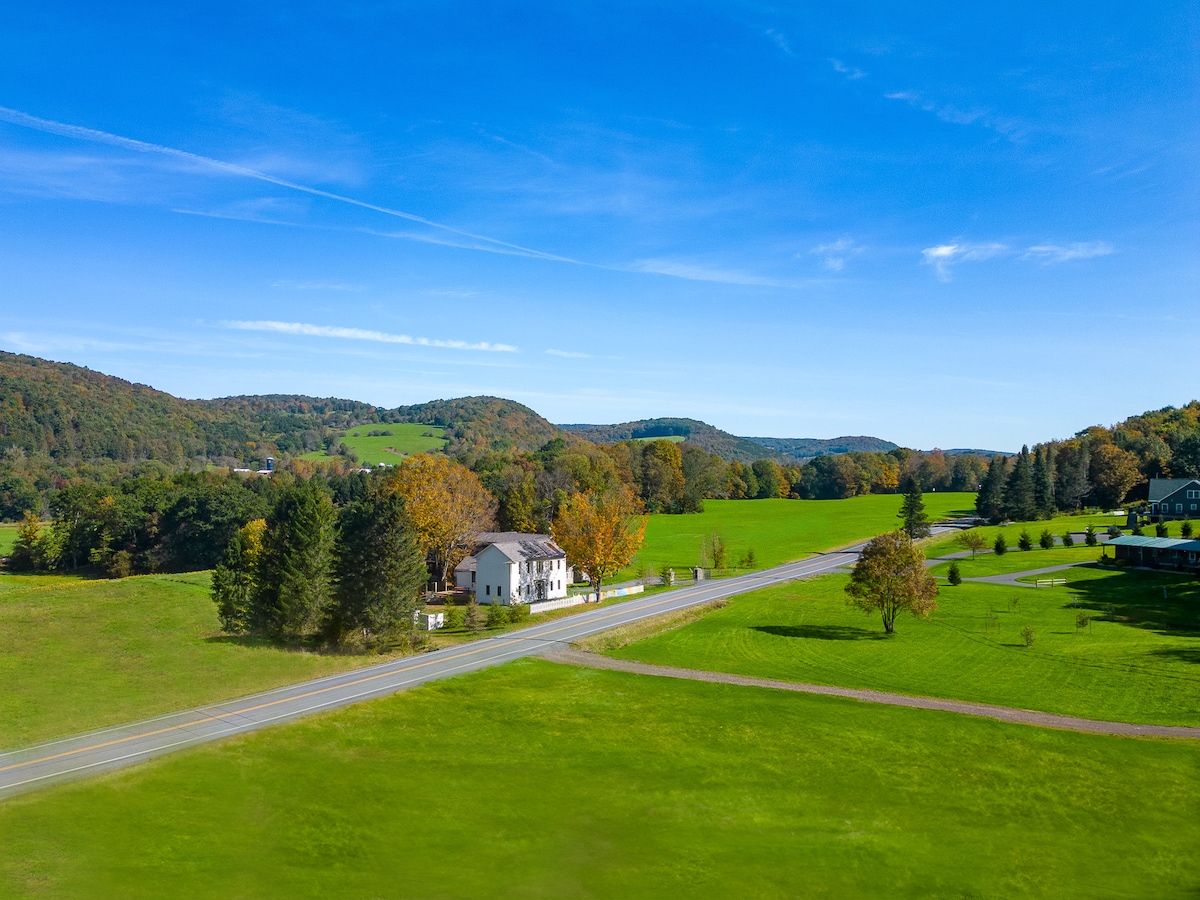 A scenic view captures a two-story home nestled beside a calm road, surrounded by lush green fields and rolling hills. Trees showcase autumn colors, while a clear blue sky enhances the peaceful rural landscape.