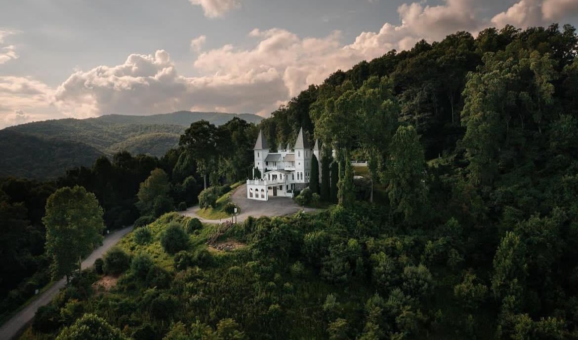 A grand castle is nestled among lush greenery and rolling hills, with its distinctive spires rising above the landscape. The scenery captures the harmony between the architectural elegance of the castle and the natural beauty of the surrounding mountains under a partly cloudy sky.