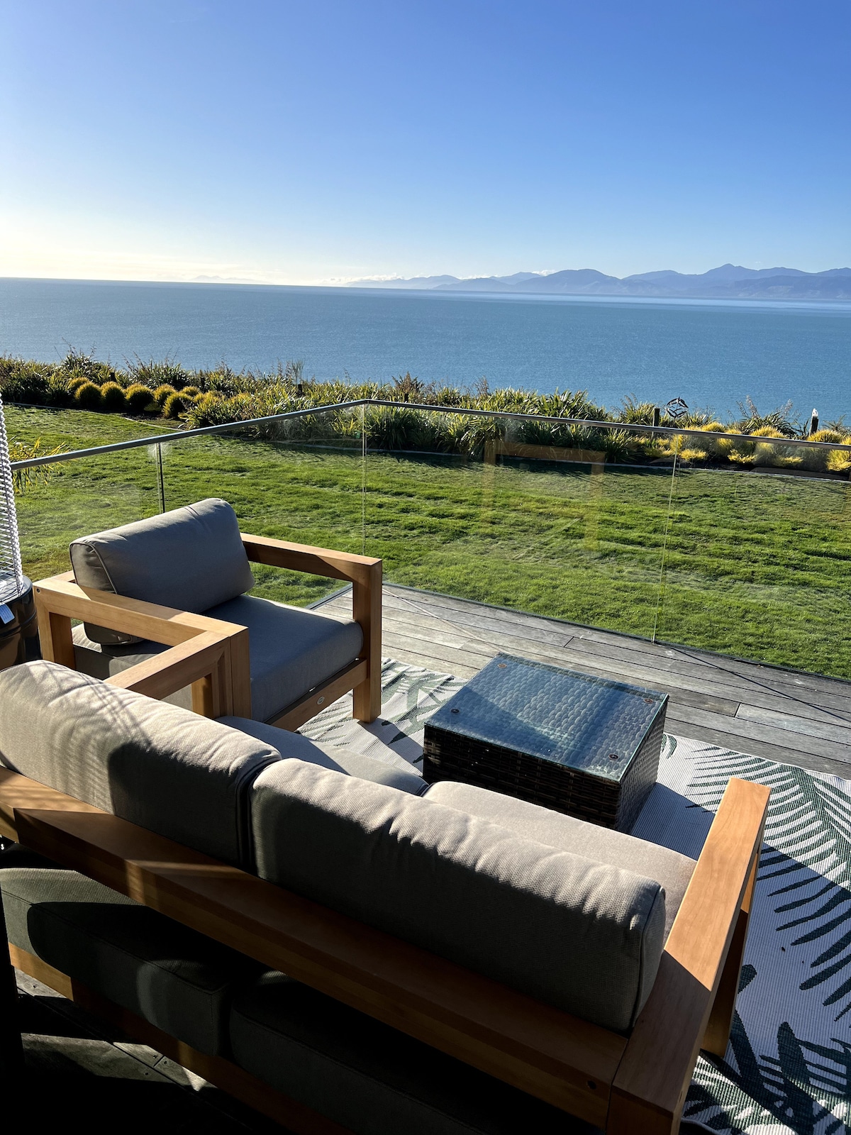 A comfortable outdoor seating arrangement features two wooden armchairs and a cushioned sofa, positioned on a deck overlooking the expansive view of Tasman Bay. A glass railing enhances the scenic vista, while greenery and coastal mountains form a picturesque backdrop.
