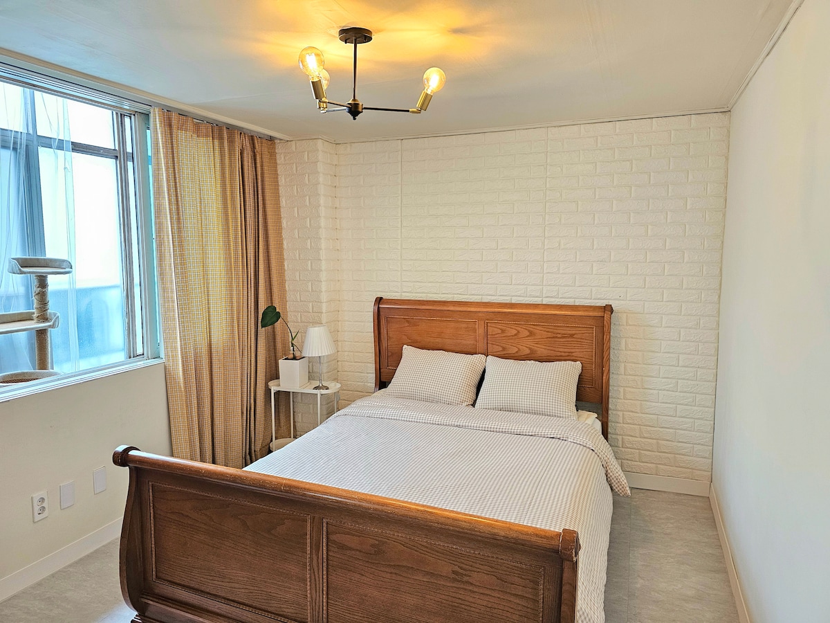 A spacious bedroom is showcased, featuring a wooden sleigh bed dressed in neutral-toned linens. Soft natural light filters through a large window adorned with sheer curtains. A bedside table with a lamp complements the minimalistic decor, while a cat tree is positioned nearby.