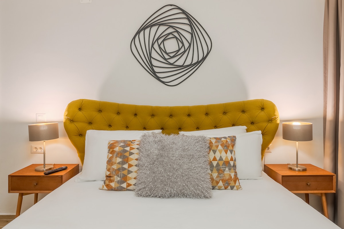 A cozy bedroom features a queen-size upholstered bed with a yellow tufted headboard. Artistic wall decor above the bed adds a modern touch. Two bedside tables, each with a lamp, provide soft lighting, creating an inviting space complemented by neutral colored bedding and decorative pillows.