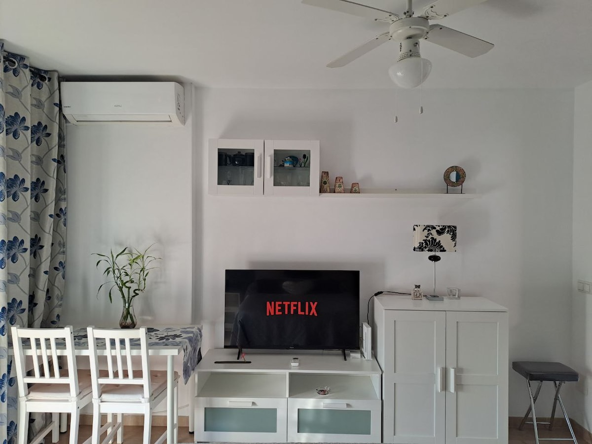A living area is depicted featuring a wall-mounted television displaying the Netflix logo. A small dining table with white chairs is positioned nearby. A white cabinet stands against the wall, accompanied by a decorative lamp. A ceiling fan provides ventilation in the bright space.