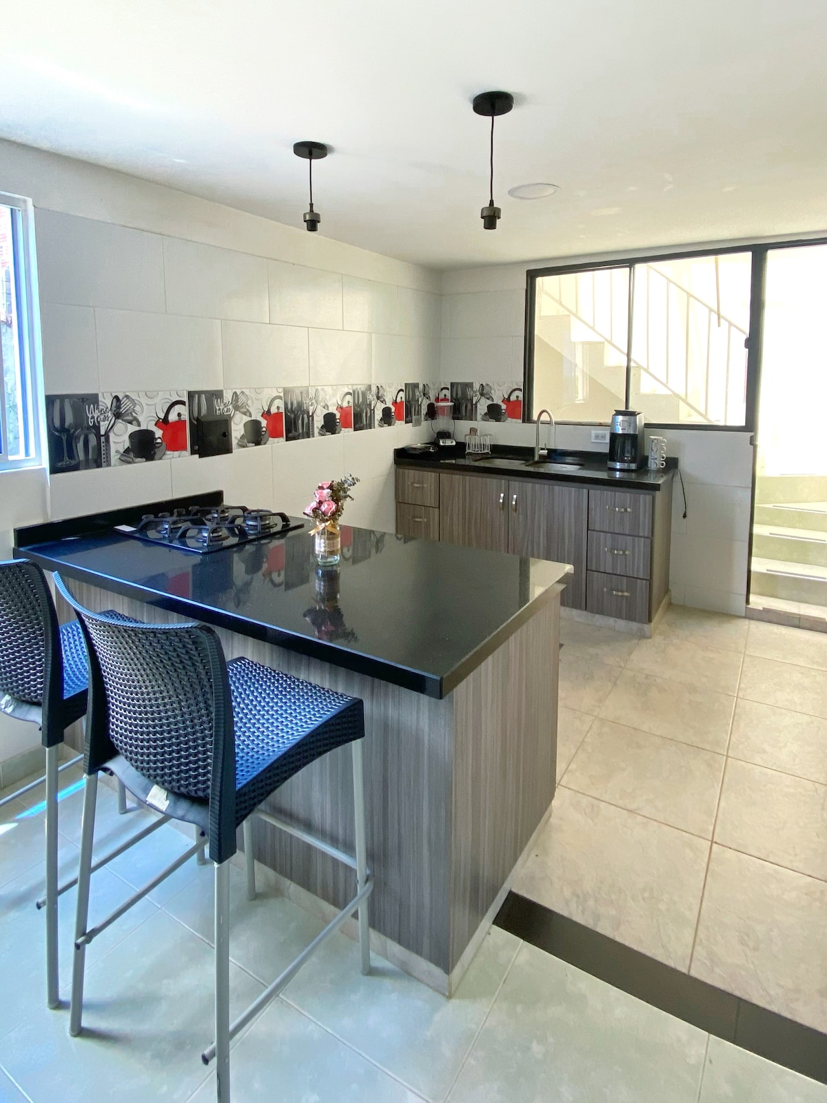 A modern kitchen features sleek cabinetry and a black countertop with seating for two. Natural light enters through a large window, illuminating the space. A coffee maker and other appliances are arranged on the counter, providing convenience for meal preparation.