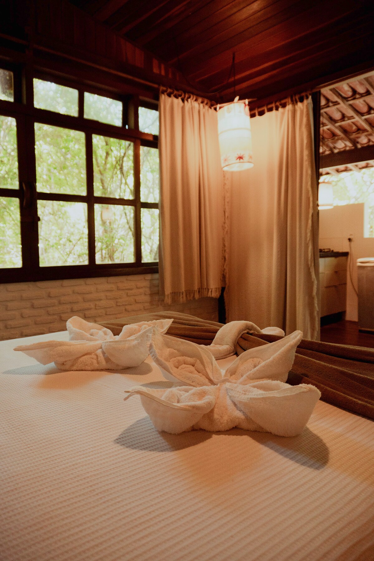 A private suite features a spacious bed adorned with elegantly arranged towel art. Natural light filters through multiple windows, surrounded by lush greenery. Soft curtains frame the windows, adding a calming touch to the tranquil atmosphere. A well-equipped kitchen is partially visible in the background.