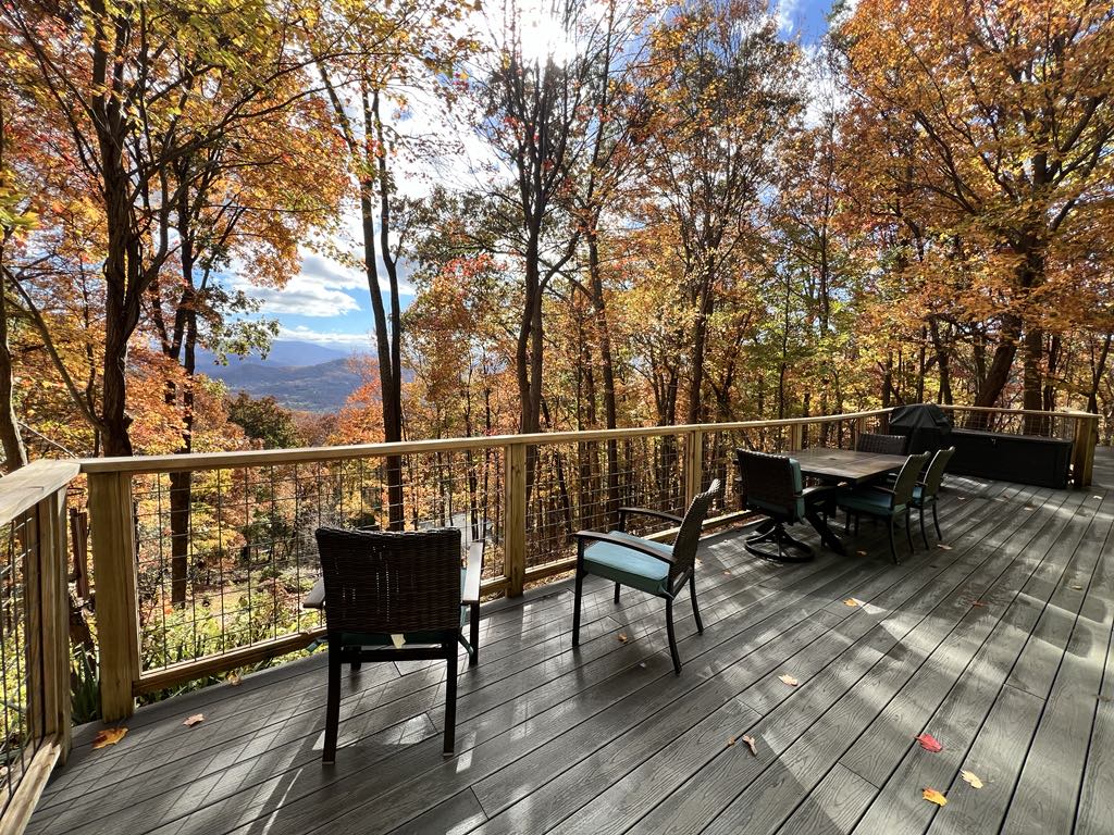 A spacious deck is surrounded by vibrant autumn foliage, offering views of distant mountains. Several chairs invite relaxation, while a table remains in the center, ideal for outdoor gatherings. Warm sunlight filters through the trees, enhancing the tranquil outdoor setting.