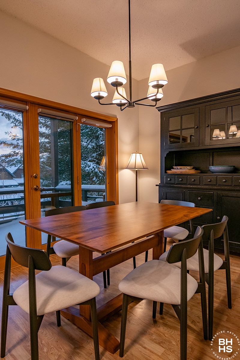 A wooden dining table is surrounded by six upholstered chairs, under a modern chandelier. The area features large glass doors that open to a covered deck, with a view of the outdoors illuminated by soft lighting. A dark cabinet is positioned against the wall.