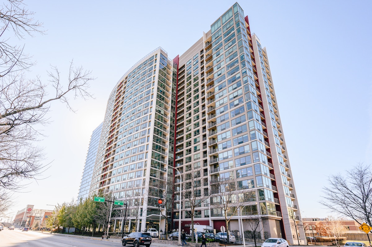 A modern high-rise building showcases a sleek façade of glass and steel, reflecting the surrounding urban environment. The structure features multiple floors with large windows, providing ample natural light. Trees line the street, contributing to the vibrant city atmosphere.