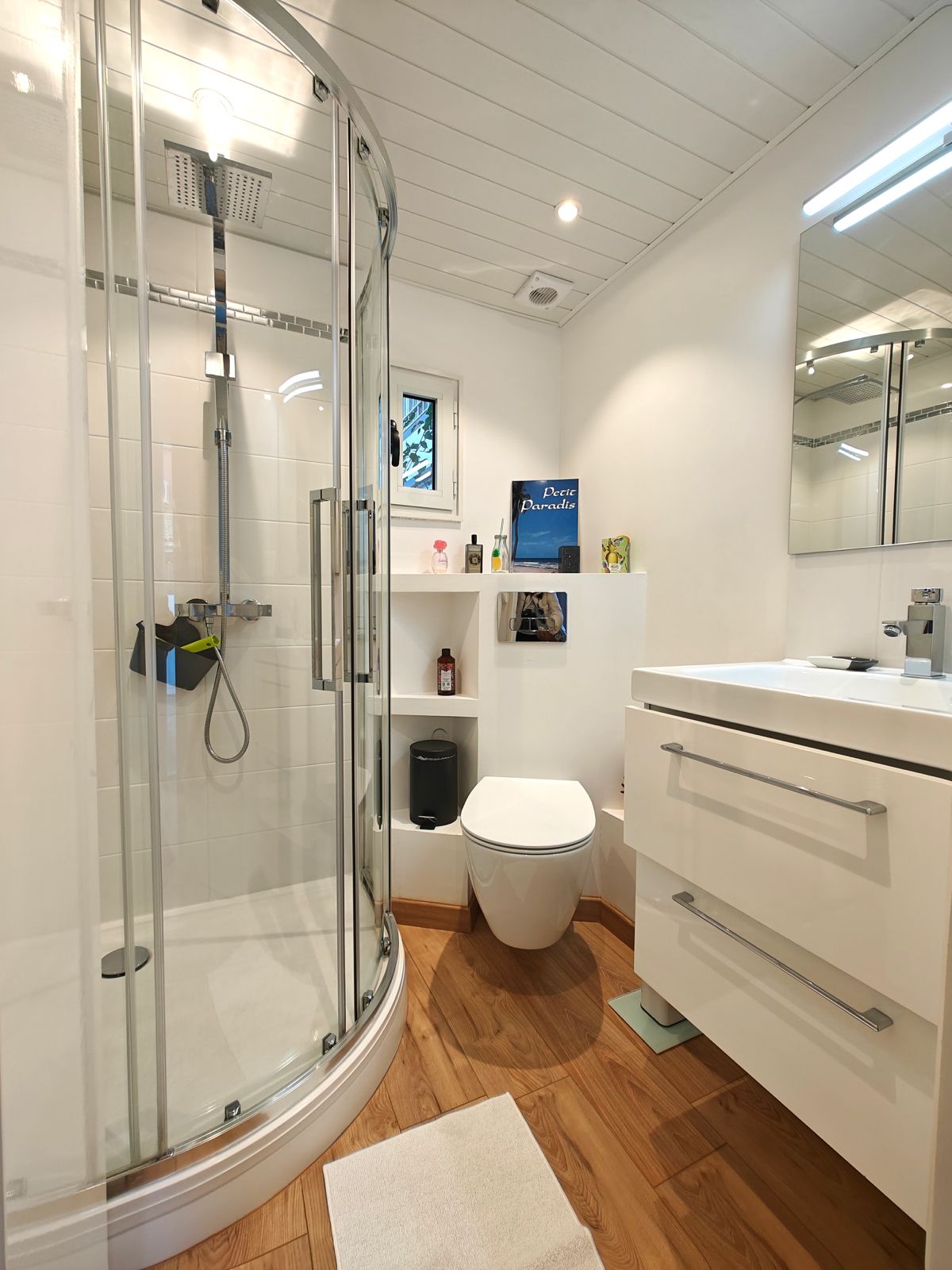 A modern bathroom features a glass-enclosed shower with a rain showerhead, a wall-mounted mirror above a white vanity with storage space, and a contemporary toilet. The warm wood flooring provides a soft contrast to the bright white tiles, complemented by neatly arranged toiletries on the shelves.