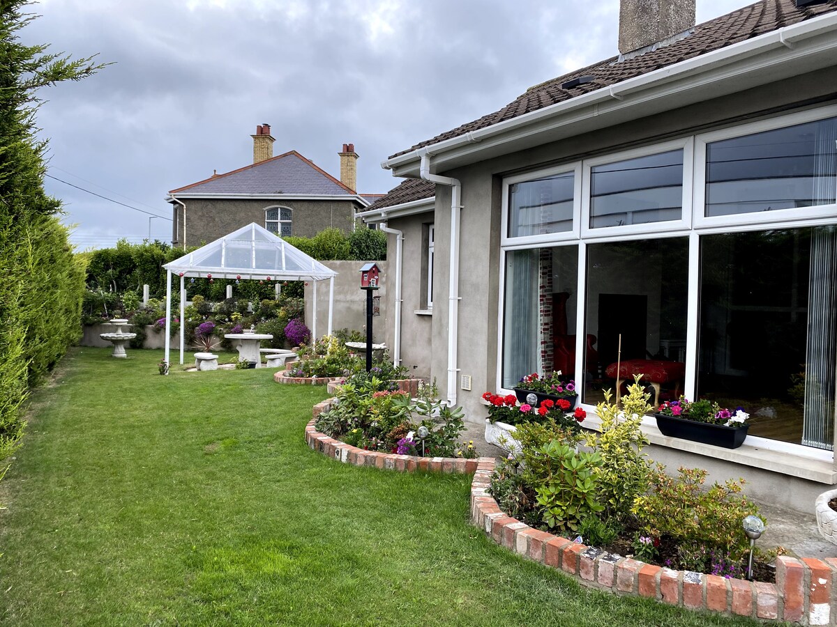 A well-maintained garden surrounds the exterior of the property, featuring colorful flower beds and manicured grass. A tent structure provides shaded outdoor space, while decorative features such as birdbaths add charm to the serene atmosphere. Large windows of the house offer views into the living area.
