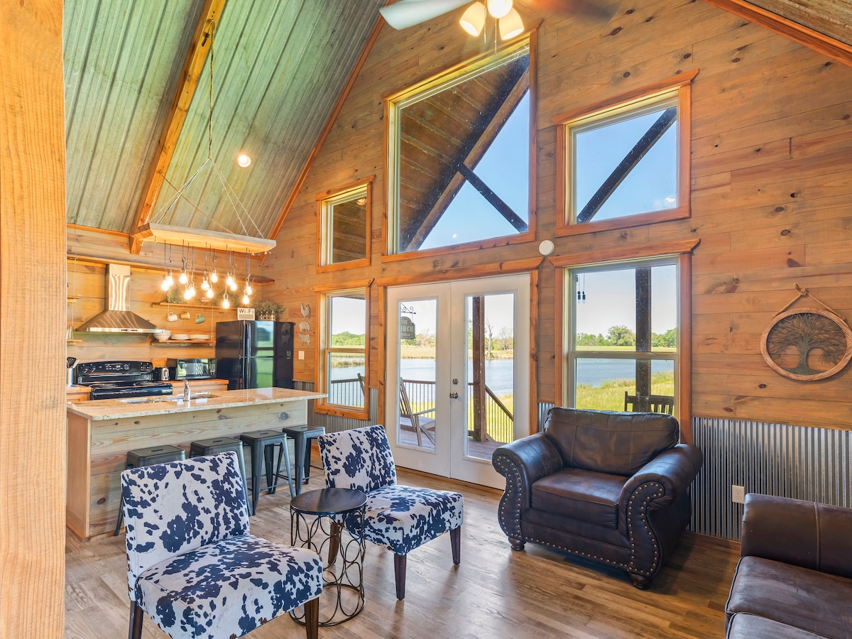 A cabin interior features a spacious living area with large windows offering views of the lake. Two patterned chairs and a leather couch provide comfortable seating, while a modern kitchen with an island and pendant lighting is visible in the background.