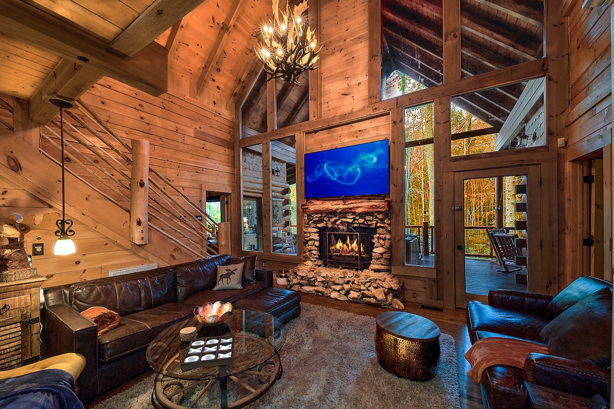 A spacious living area features a stone fireplace beneath a mounted television, surrounded by large windows offering views of the forest. Leather seating arrangements are present, with a central coffee table and a comfortable rug beneath. Warm wood tones create a cozy ambiance.