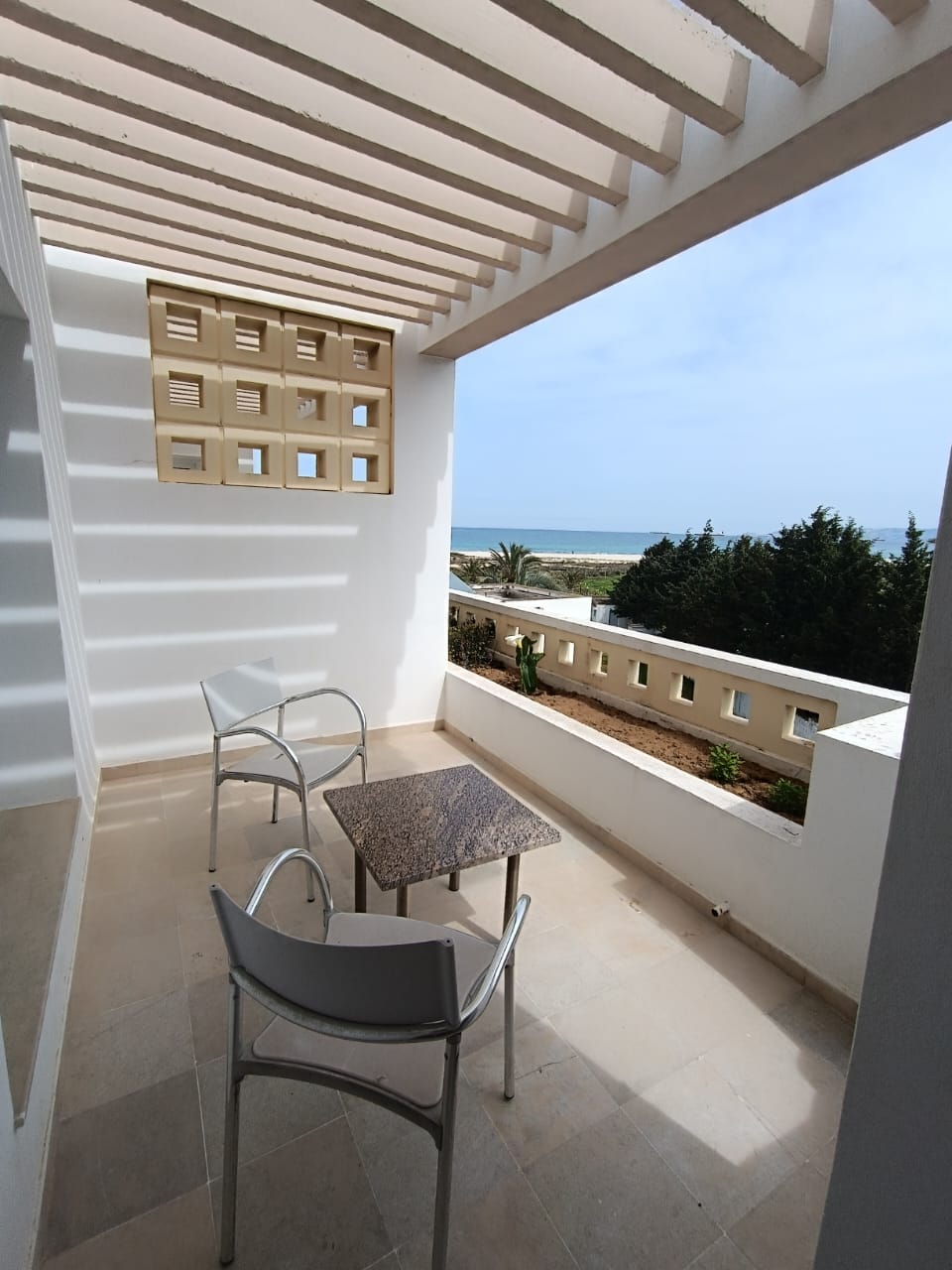 A small balcony features two simple white chairs and a square table made of dark stone. The design incorporates striking white walls and a decorative element with cut-out patterns. A serene view of the sea and greenery is visible in the background.