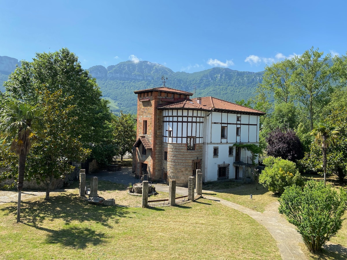 The exterior of a historic building is visible, featuring a mix of stone and wooden elements. Lush greenery surrounds the structure, with trees and manicured grass creating a serene backdrop. Nearby mountains are visible against a clear blue sky, enhancing the natural landscape.