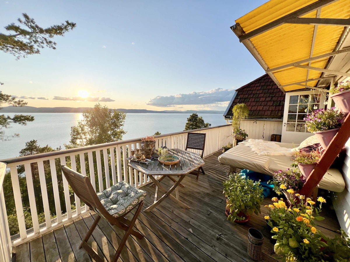 A spacious terrace overlooks the sea, bathed in warm sunset light. A wooden table is set for dining, accompanied by potted flowers and greenery. Comfortable seating is positioned nearby, enhancing the peaceful outdoor ambiance.