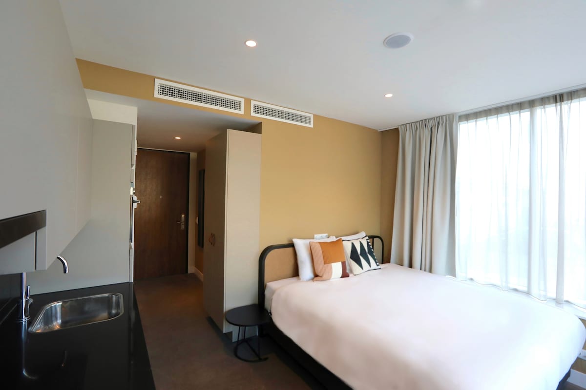 A spacious studio features a comfortable bed with neutral bedding. A kitchenette with a small sink is positioned alongside a minimalist table and chair. Natural light fills the room through large windows adorned with sheer curtains, and warm tones create an inviting atmosphere.