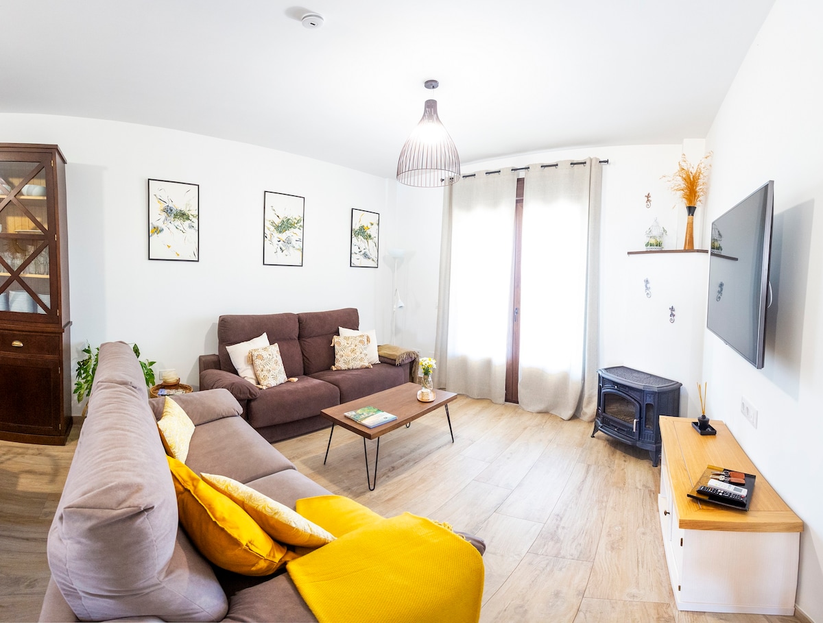 A cozy living area is arranged with a dark brown sofa and a light gray sectional, complemented by decorative cushions. Artwork with floral themes adorns the walls. A flat-screen TV is mounted across from the seating, with a small wooden table and a decorative fireplace nearby.