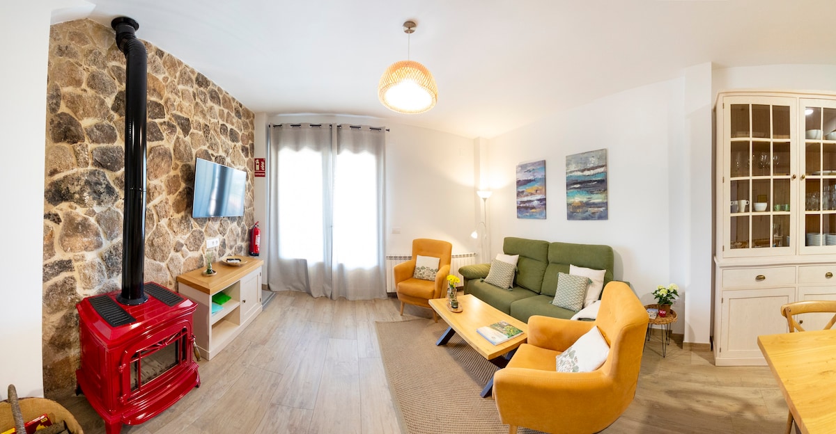 A cozy living room features a charming stone wall and a vibrant red wood stove. A green sofa and two orange armchairs are positioned around a wooden coffee table. Artwork adorns the walls, and natural light enters through a large window covered by light curtains.