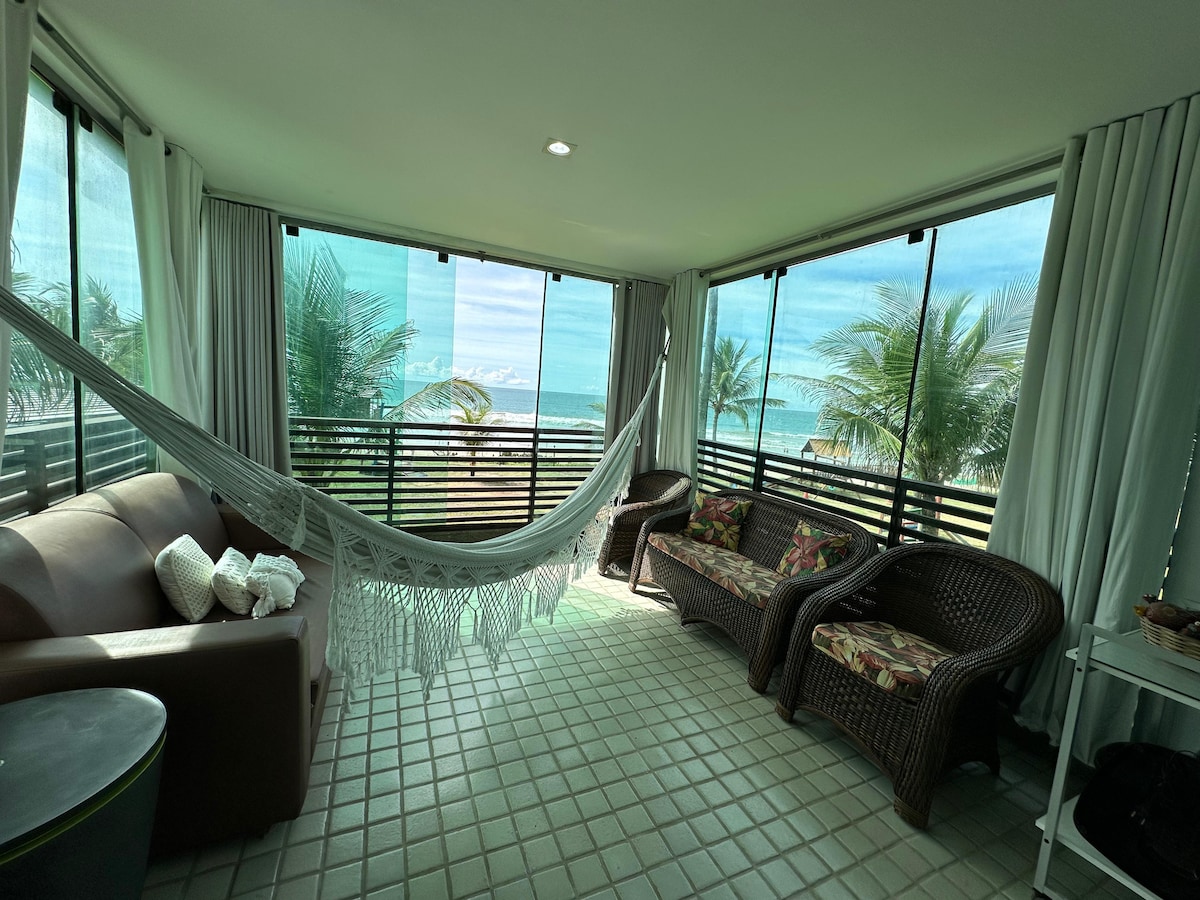 A cozy balcony space offers views of palm trees and the ocean. Seating includes a sofa and two wicker chairs adorned with colorful cushions. A hammock is suspended between posts, adding to the inviting atmosphere. Large glass windows allow natural light to fill the room.