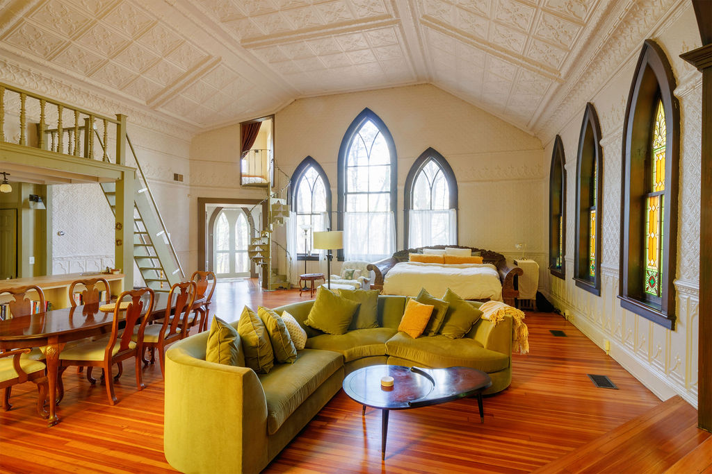 The spacious great room is characterized by 25-foot cathedral ceilings and original stained glass windows. A cozy sectional sofa in a soothing green hue surrounds a dark coffee table, with a queen bed positioned in a corner. Light streams in, enhancing the room's unique architectural details.
