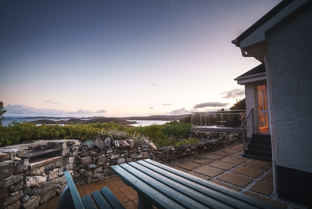 An inviting outdoor area features a stone wall and a cozy seating arrangement overlooking the expansive views of the sea and mainland. The soft colors of the sky blend with the serene landscape as twilight sets in, creating a tranquil atmosphere for relaxation.
