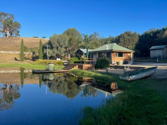 The boat house is visible beside a tranquil dam, surrounded by greenery. Steps lead from the house to the water's edge, where a small boat is moored. Sunshine reflects off the water, creating a serene atmosphere complemented by the natural landscape.