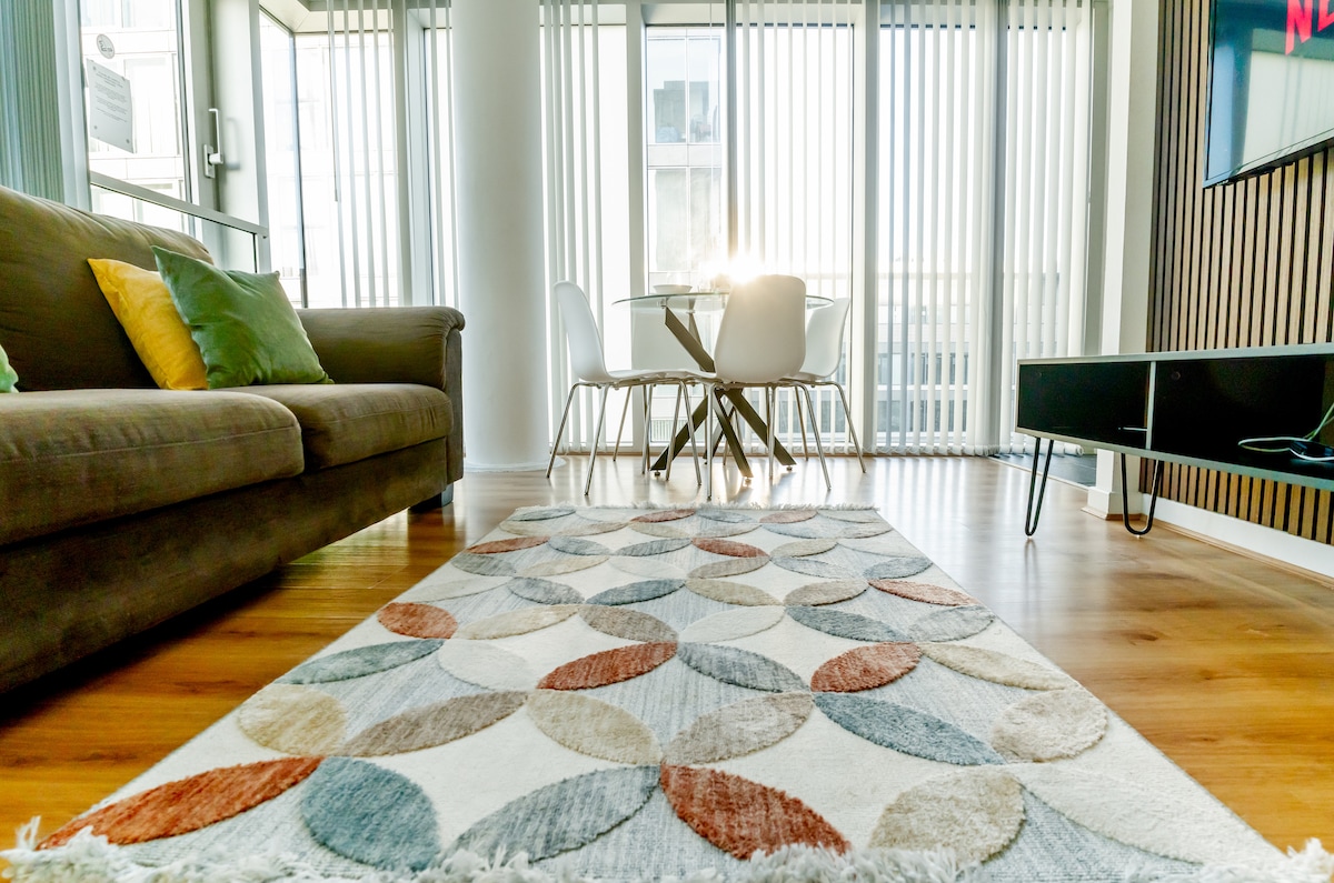 A spacious living area showcases a patterned area rug, complemented by a comfortable sofa adorned with decorative cushions. A dining table for four sits nearby, with natural light filtering through the vertical blinds, enhancing the modern decor and welcoming ambiance of the space.
