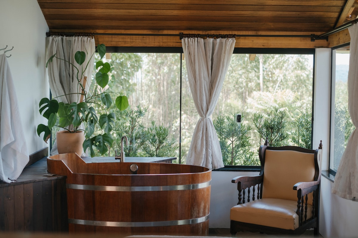 A wooden soaking tub is highlighted beside large windows that allow ample natural light, offering views of the surrounding greenery. A comfortable armchair provides a relaxing spot, while sheer curtains frame the windows, enhancing the serene atmosphere of the space.