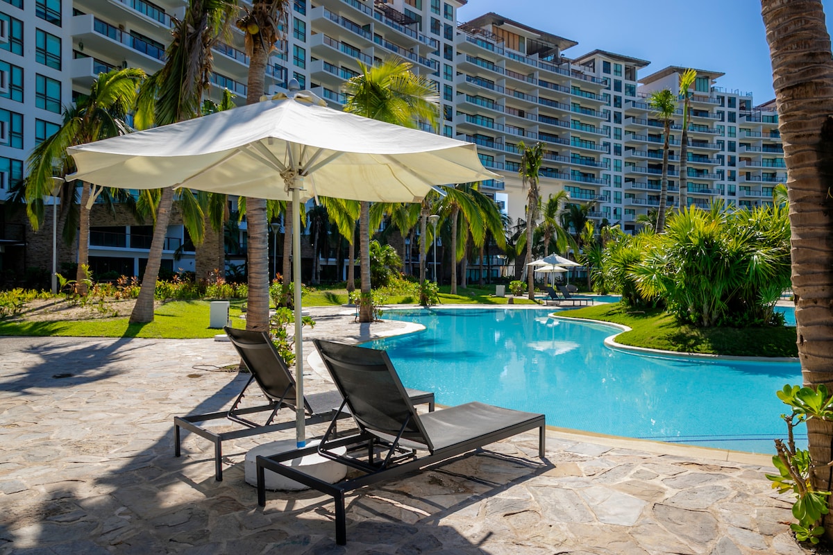 Two loungers are positioned under a large white umbrella beside a clear blue pool, surrounded by lush palm trees and tropical landscaping. The modern residential towers are visible in the background, complementing the serene outdoor setting.