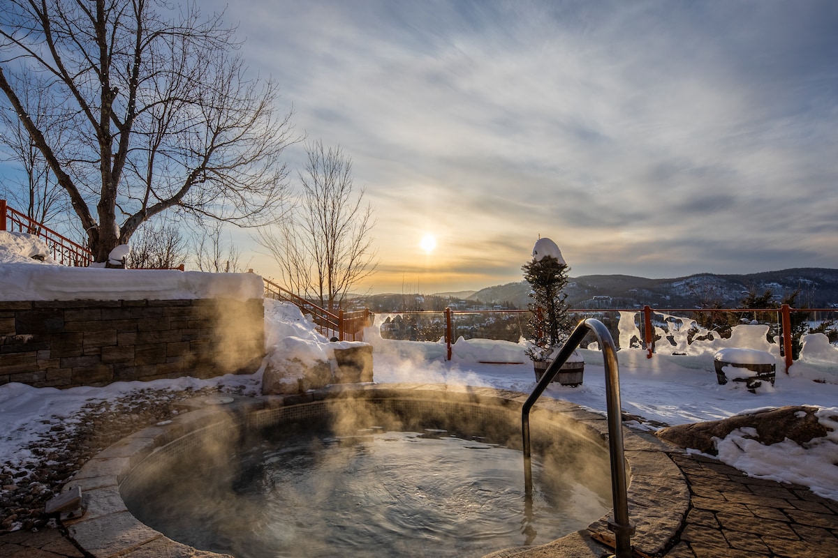 A hot tub sits amidst a snowy landscape, with steam rising gently into the crisp air. The setting sun casts warm hues across the sky, while distant hills and trees provide a serene backdrop. Outdoor seating is visible on the terrace nearby.