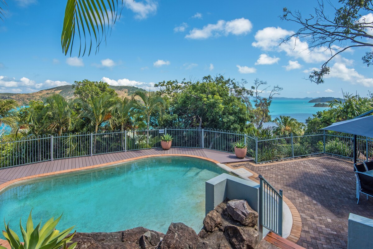 A private pool is set against a backdrop of lush greenery and expansive ocean views. A wooden deck surrounds the pool, complemented by decorative planters. The sky features scattered clouds and bright blue tones, enhancing the serene outdoor space inviting relaxation.