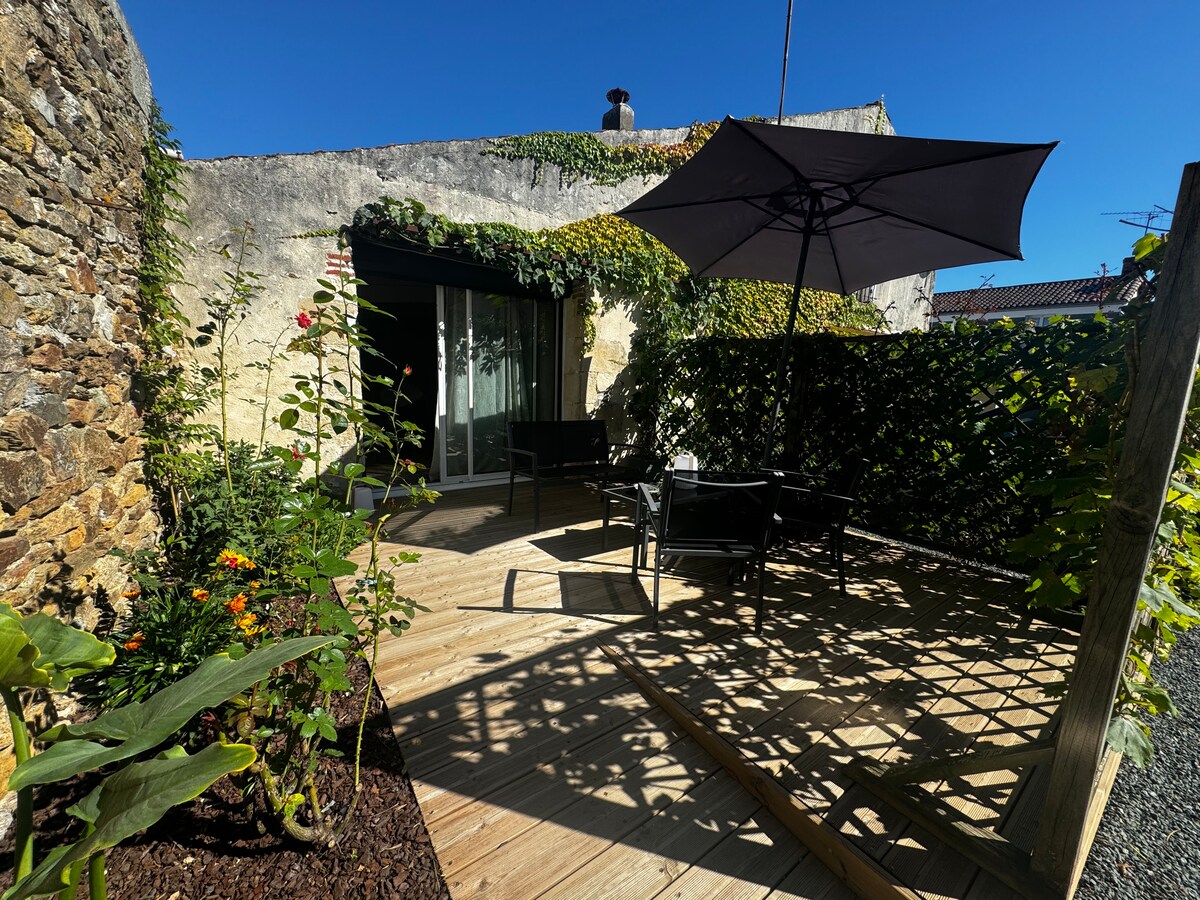 A private outdoor terrace is surrounded by lush greenery and flowers. Comfortable black seating is shaded by a large umbrella. Natural stone walls feature climbing plants, and sunlight filters through the leaves, creating a peaceful atmosphere.
