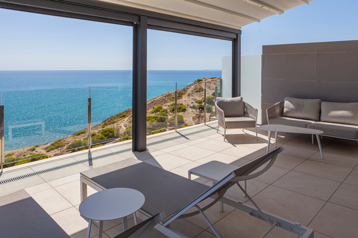 An outdoor terrace features modern seating arrangements and sun loungers, providing sweeping views of the Mediterranean Sea. The space is designed with clear glass railings and is surrounded by light-colored tiles, emphasizing the tranquil atmosphere and inviting natural light.