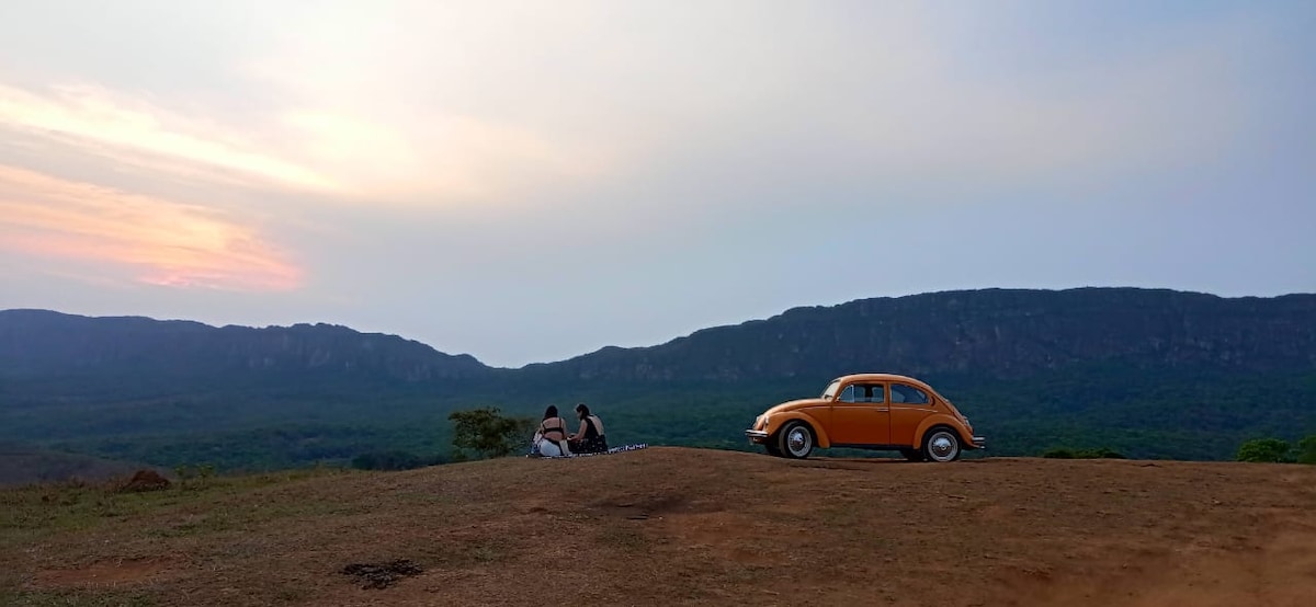An orange vintage car is parked on a grassy hill, overlooking a vast expanse of green wooded land. Two individuals are seated on the ground, facing a distant mountain range under a softly lit sky during sunset.