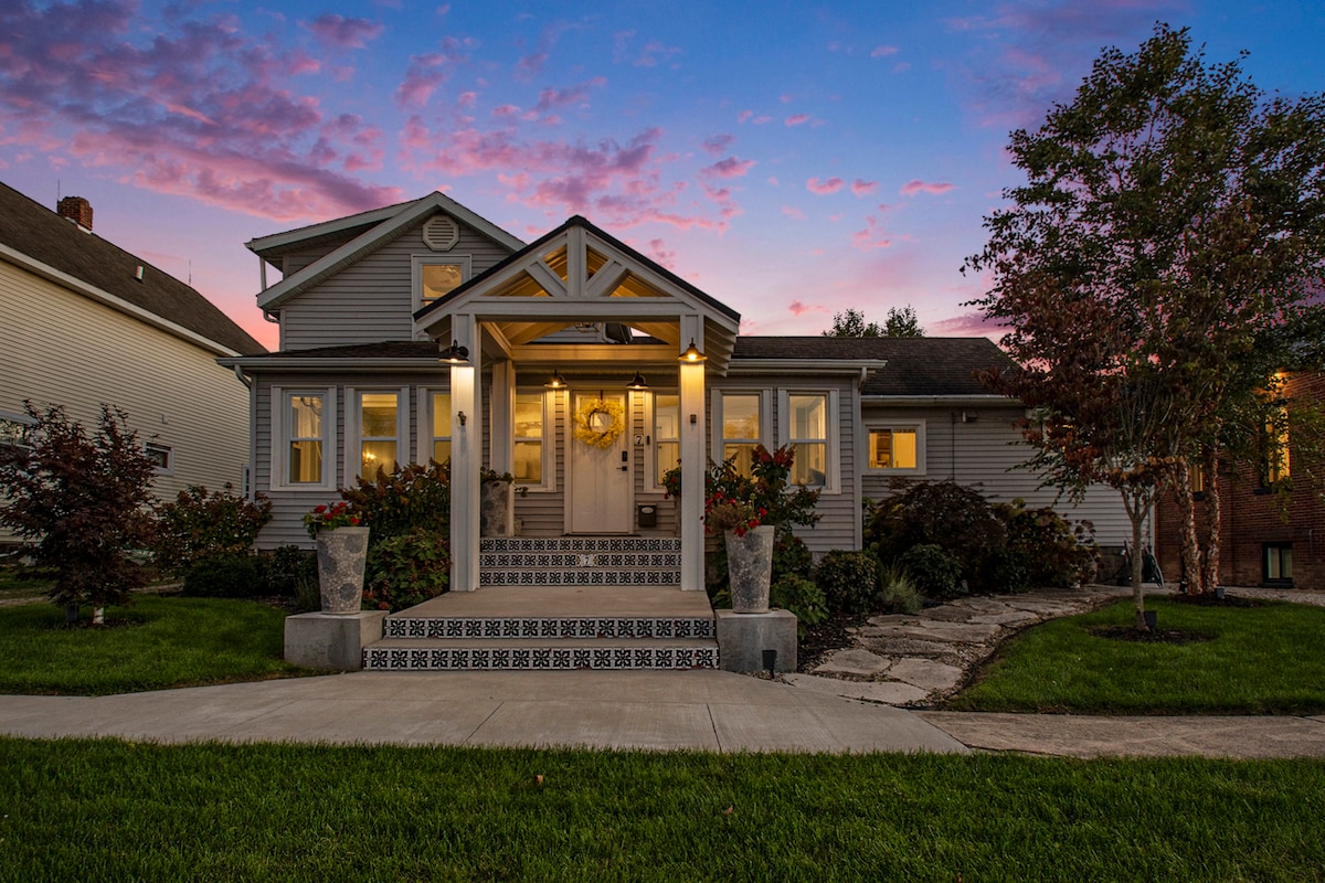 An updated home is positioned at the center of the image, featuring a welcoming entrance with a decorative porch and large front steps. Surrounding gardens include well-maintained shrubs and trees. The sky showcases soft pink and purple hues as daylight transitions into evening.