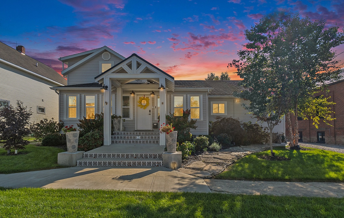 The exterior of a charming updated home is presented, featuring a welcoming porch with a decorative sun emblem. Lush green grass and flower beds frame the entrance, while the vibrant sky showcases hues of purple and orange, hinting at a picturesque sunset.