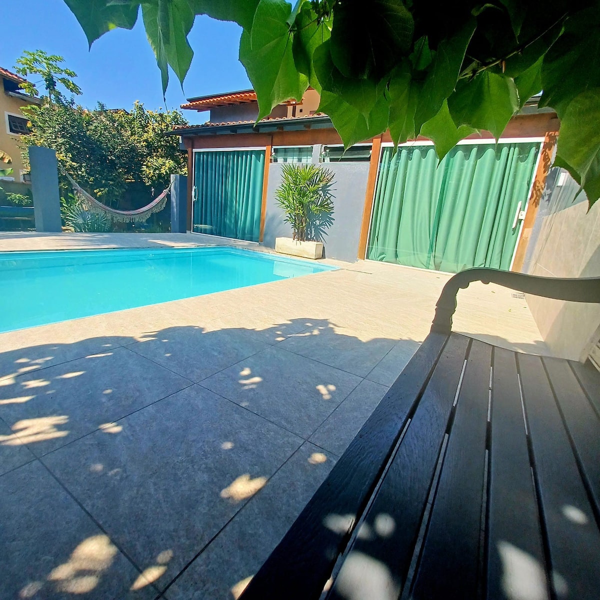 A serene outdoor space features a refreshing pool surrounded by tiled flooring. Lush greenery provides shade, while a hammock is visible nearby. Large green curtains frame the entrance to adjacent suites, enhancing the tranquil atmosphere of the area.