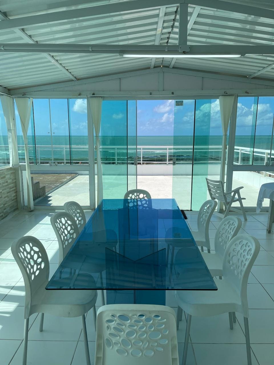 A bright and airy dining area features a large glass table surrounded by white chairs, all set against sweeping views of the ocean through large floor-to-ceiling windows. The space is illuminated by natural light, creating a relaxing atmosphere.