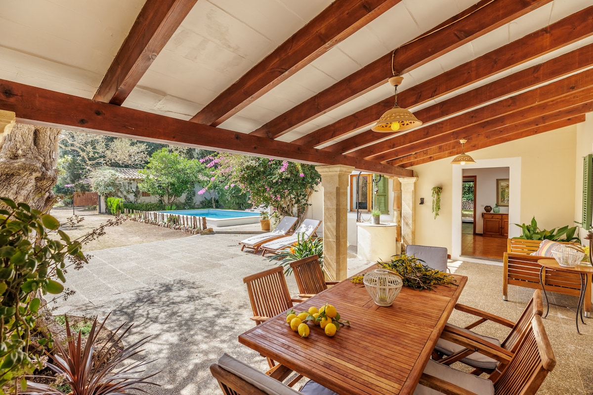 An outdoor dining area is shown with a wooden table and comfortable chairs, shaded under a pergola with wooden beams. Lush green landscaping surrounds the space, and a glimpse of the pool area can be seen in the background, inviting relaxation.