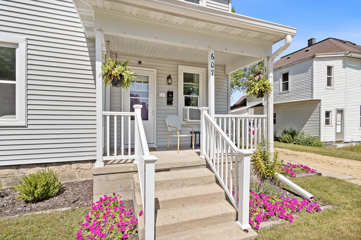 The entrance features a welcoming porch with white railings and steps leading to a covered entryway. Potted plants hang from the ceiling, and a pair of chairs are placed on the porch, with colorful flowers lining the path leading to the door.