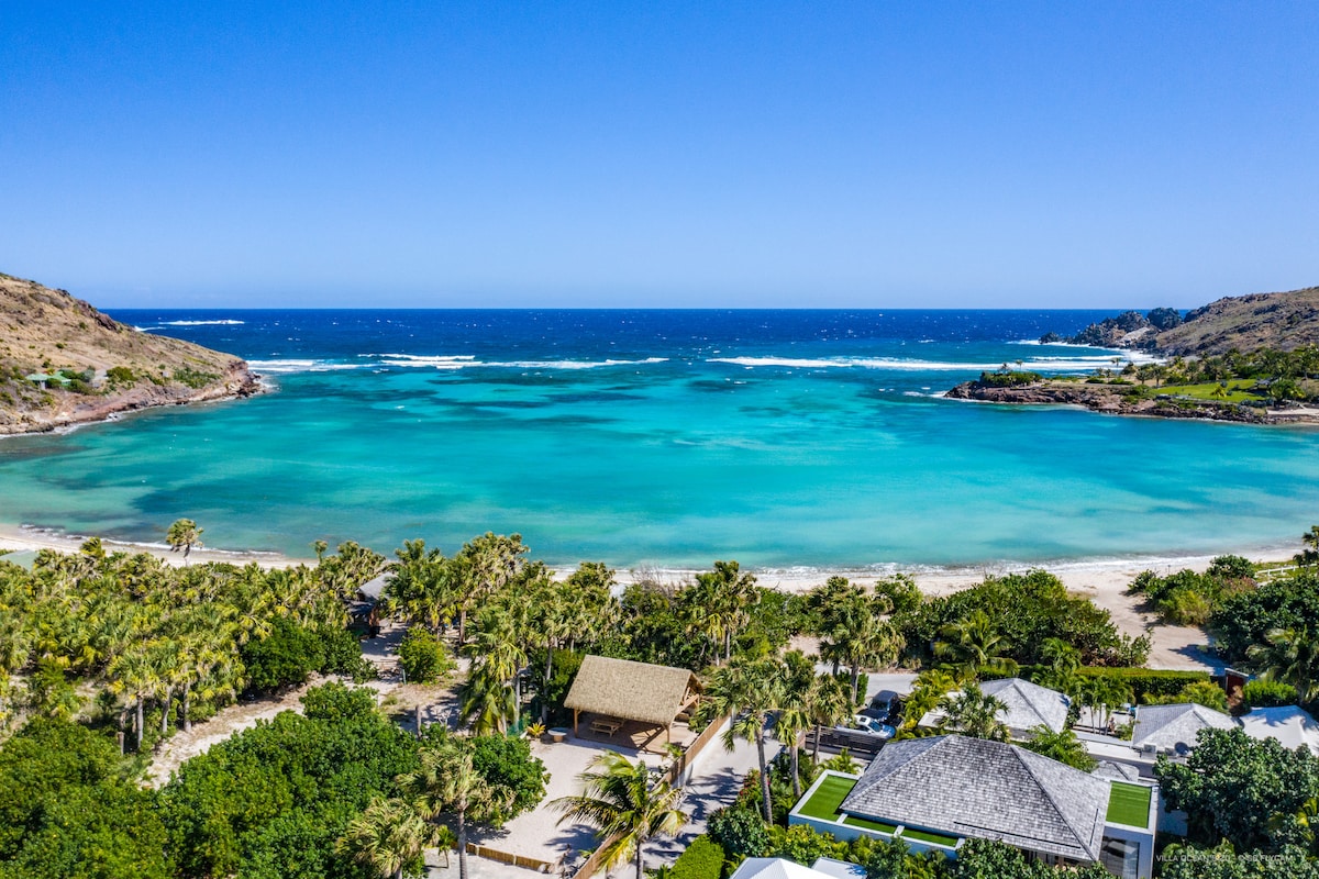 An aerial view captures the tranquil turquoise lagoon bordered by lush greenery and sandy shores. Palm trees punctuate the landscape, complementing the calm waves lapping against the beach, with distant hills framing the serene coastal scene.
